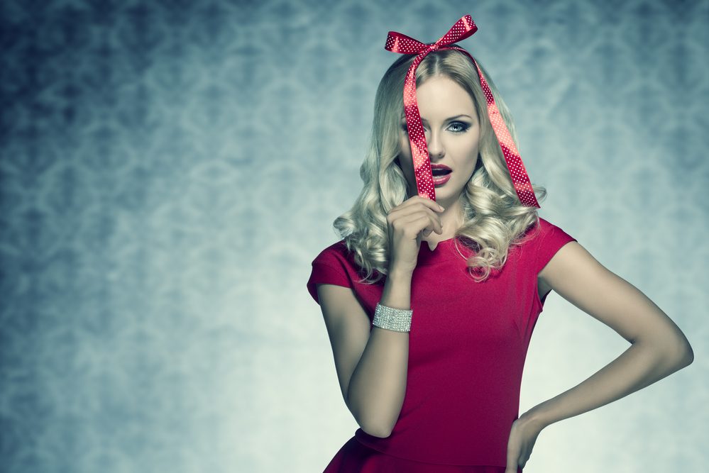 christmas shoot of beauty blonde elegant woman with red fashion dress and bright bracelet posing with funny bow on her head like a present