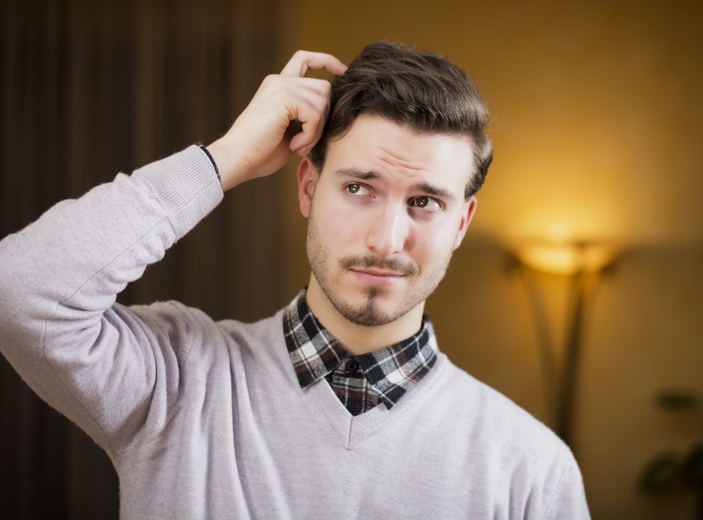 Confused or doubtful young man scratching his head and looking up. Indoors shot in a living room