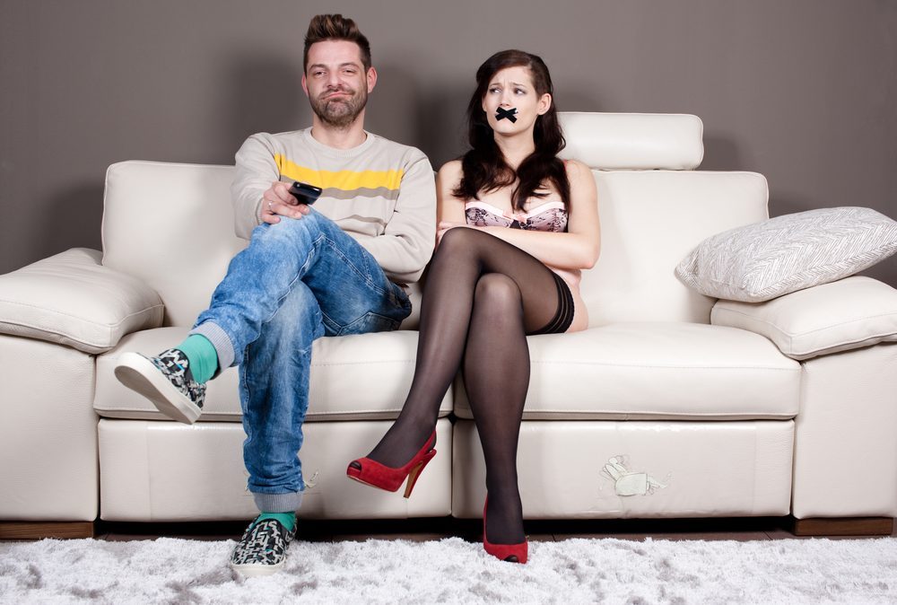 I finally managed to shut her up! Young woman with taped mouth sitting beside her boyfriend/husband who's enjoying in silence and watching TV.