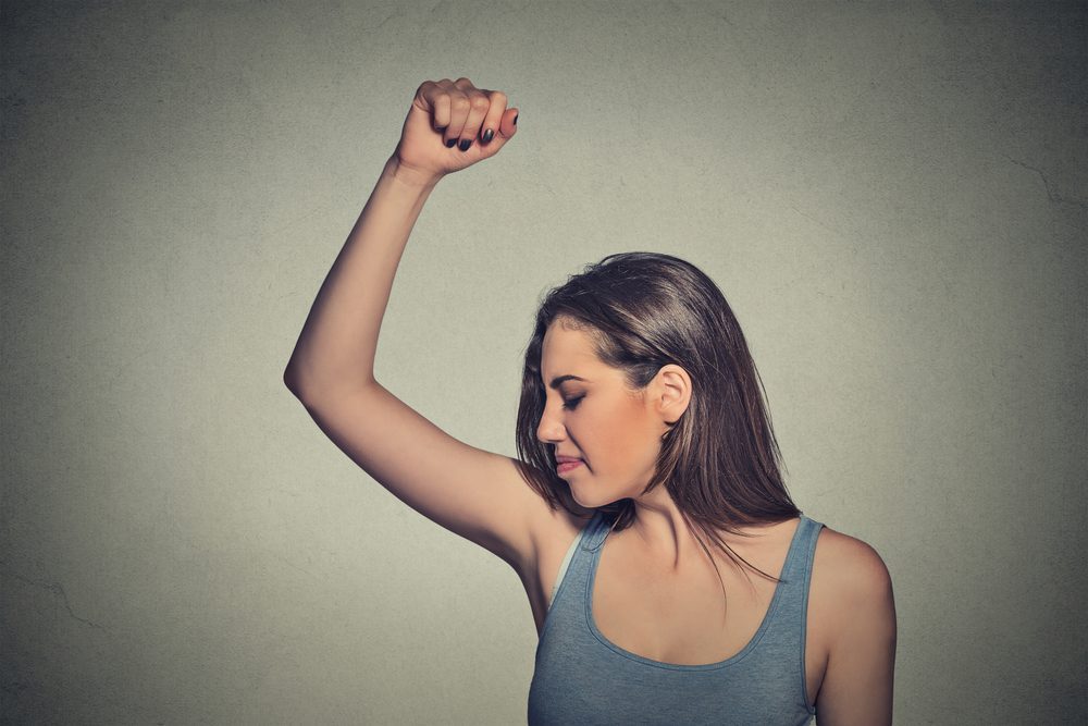 Closeup portrait young woman, smelling, sniffing her wet armpit, something stinks, very bad foul odor situation isolated grey wall background. Negative human emotion facial expression feeling reaction