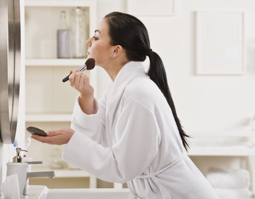 A young woman is putting on makeup in front of a mirror. She is looking away from the camera. Horizontally framed shot.