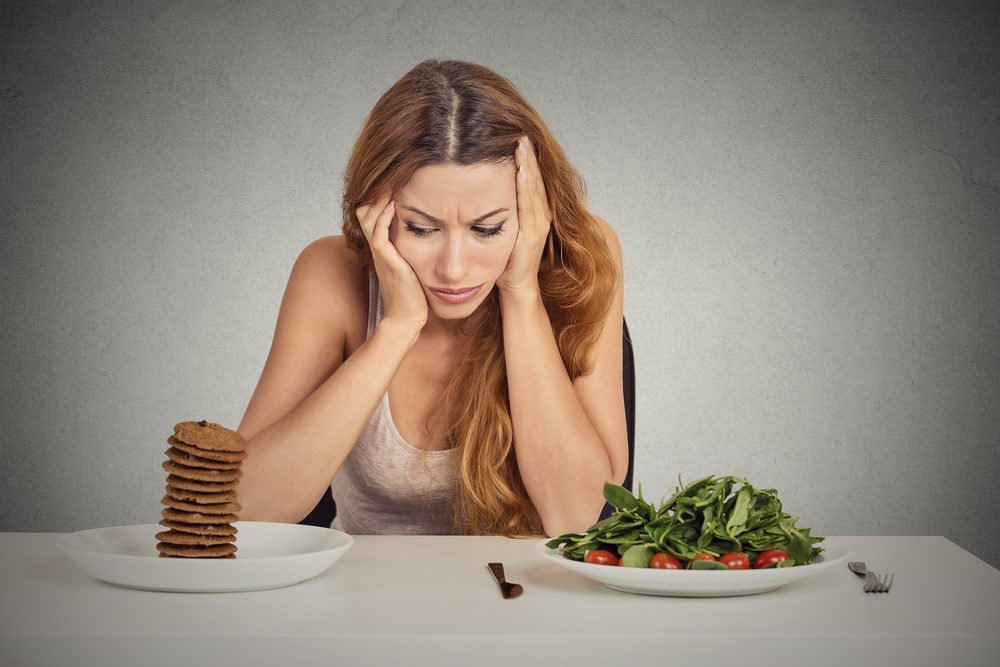 Young woman tired of diet restrictions deciding whether to eat healthy food or sweet cookies she is craving sitting at table isolated grey background. Human face expression emotion. Nutrition concept