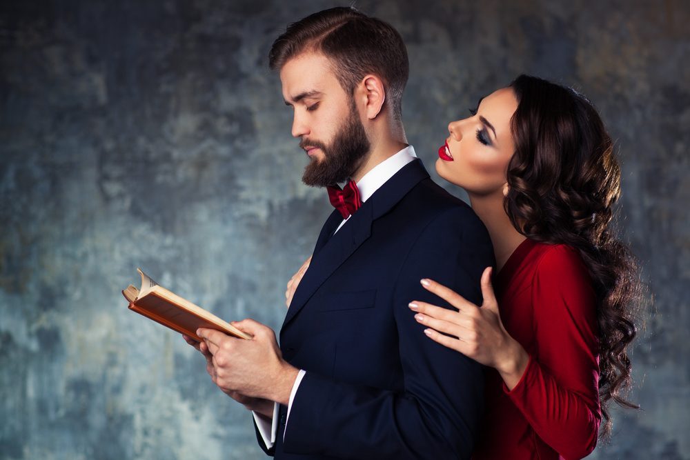 Young elegant couple in evening dress portrait. Man reading book and woman trying to attract and embrace him.