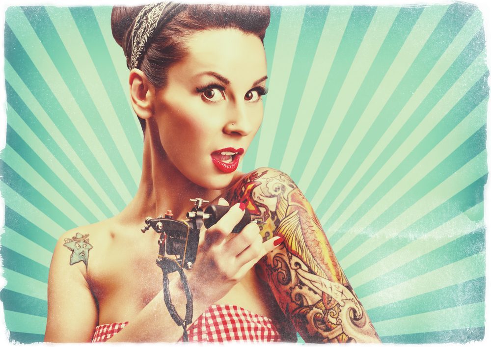 Photo of beautifull pin-up girl with tattoos and tattoo machine tattoing herself and looking at the camera