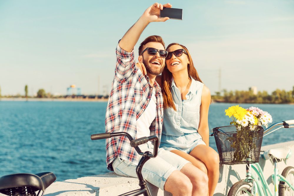 Collecting the bright moments. Smiling young couple making selfie while sitting on parapet near their bicycles