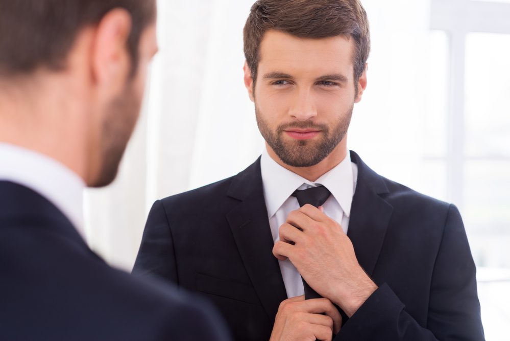 Looking just perfect. Handsome young man in formalwear adjusting his necktie and smiling while standing against mirror