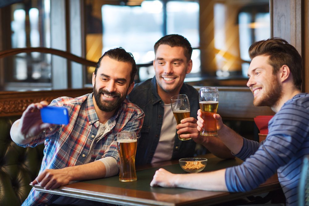 people, men, leisure, friendship and technology concept - happy male friends drinking beer and taking selfie with smartphone at bar or pub